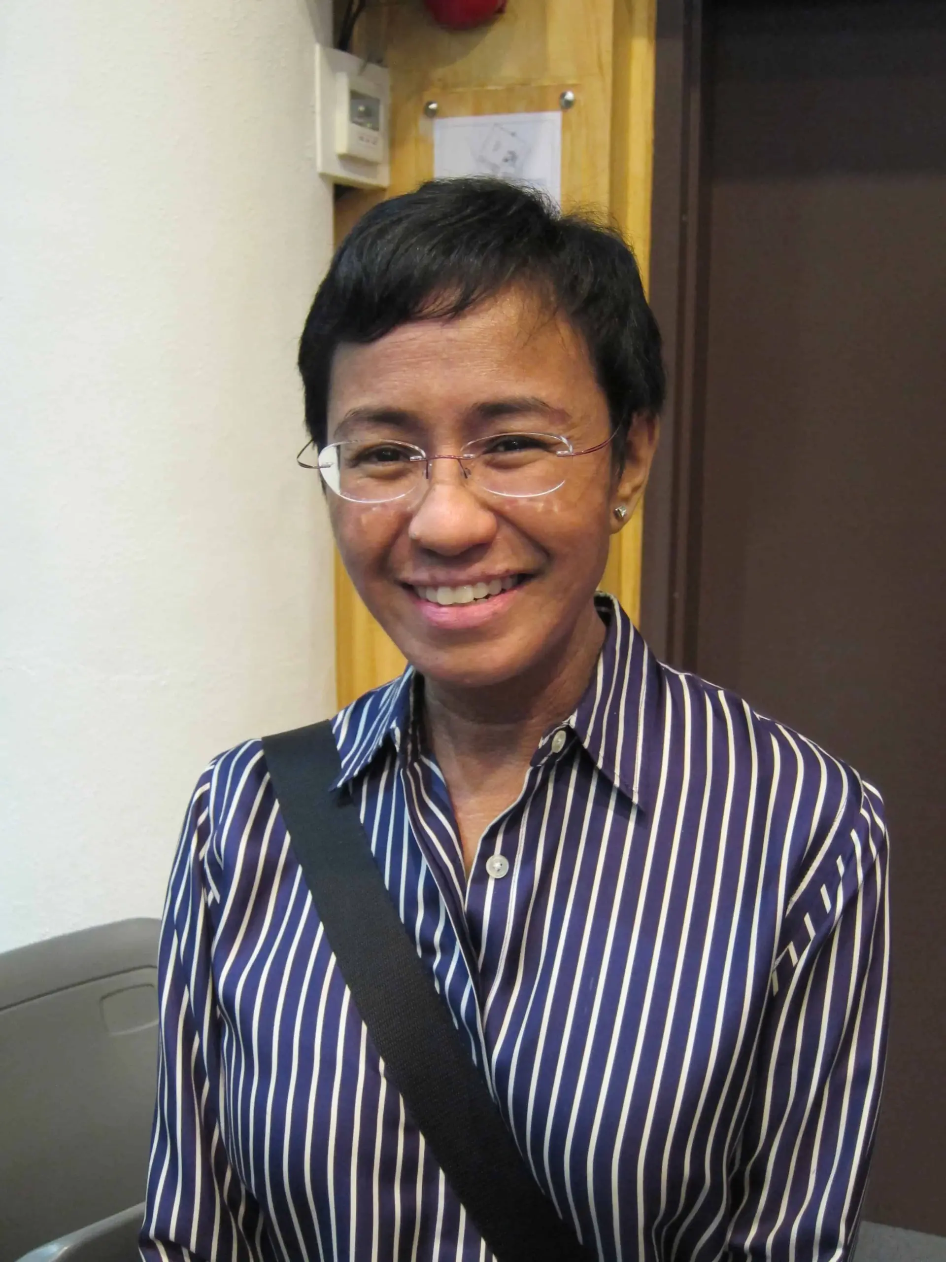 A person with short dark hair, wearing glasses and a blue-and-white striped shirt, smiles at the camera. They have a black shoulder strap across their chest and stand in an indoor setting with a wooden background.