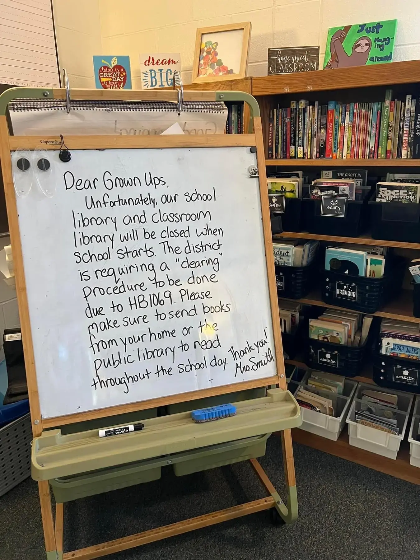 A classroom library with shelves of books and bins. A whiteboard on an easel displays a handwritten note explaining the school library is closed due to a “clearing procedure” under HB1069 and asks families to send books from home.