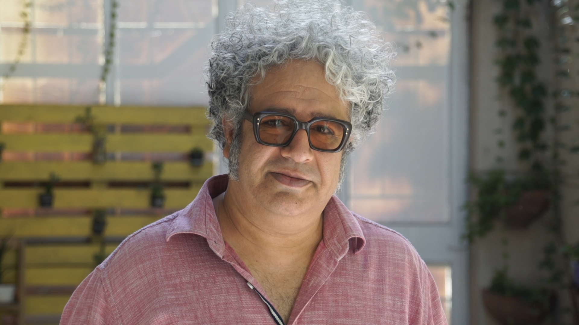 A man with curly gray hair and large glasses wears a pink shirt and stands outdoors in front of a yellow wooden structure and plants.