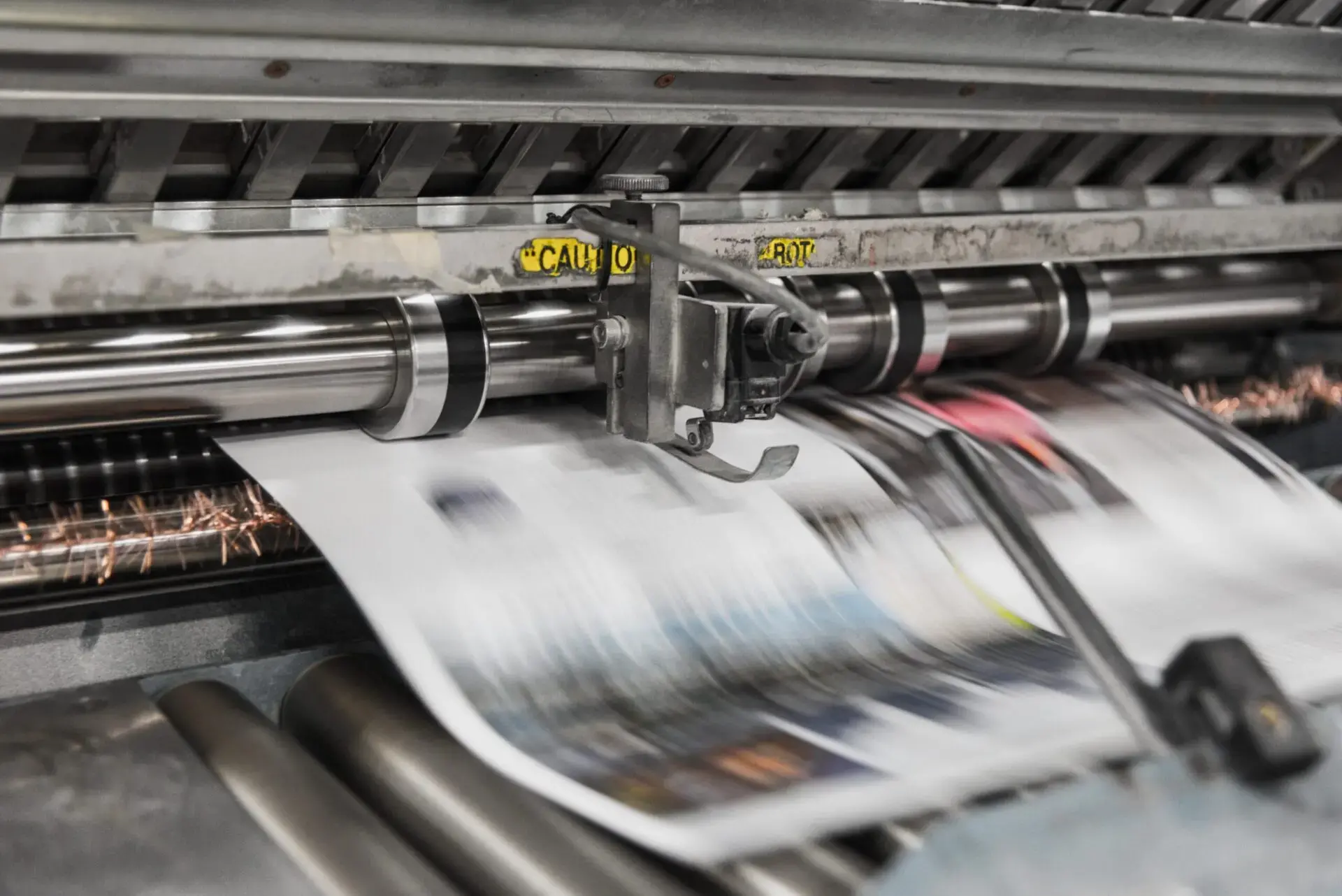 Close-up of a printing press in action, rapidly printing colorful sheets of paper. The machinery is metallic with warning labels, and the printed sheets appear blurred due to motion.