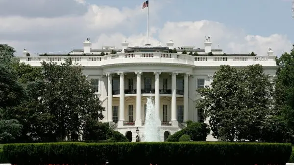 The White House, a large neoclassical building with columns, stands behind a fountain and manicured hedges, the U.S. flag flying above—a symbol of free press sometimes threatened, under a partly cloudy sky.