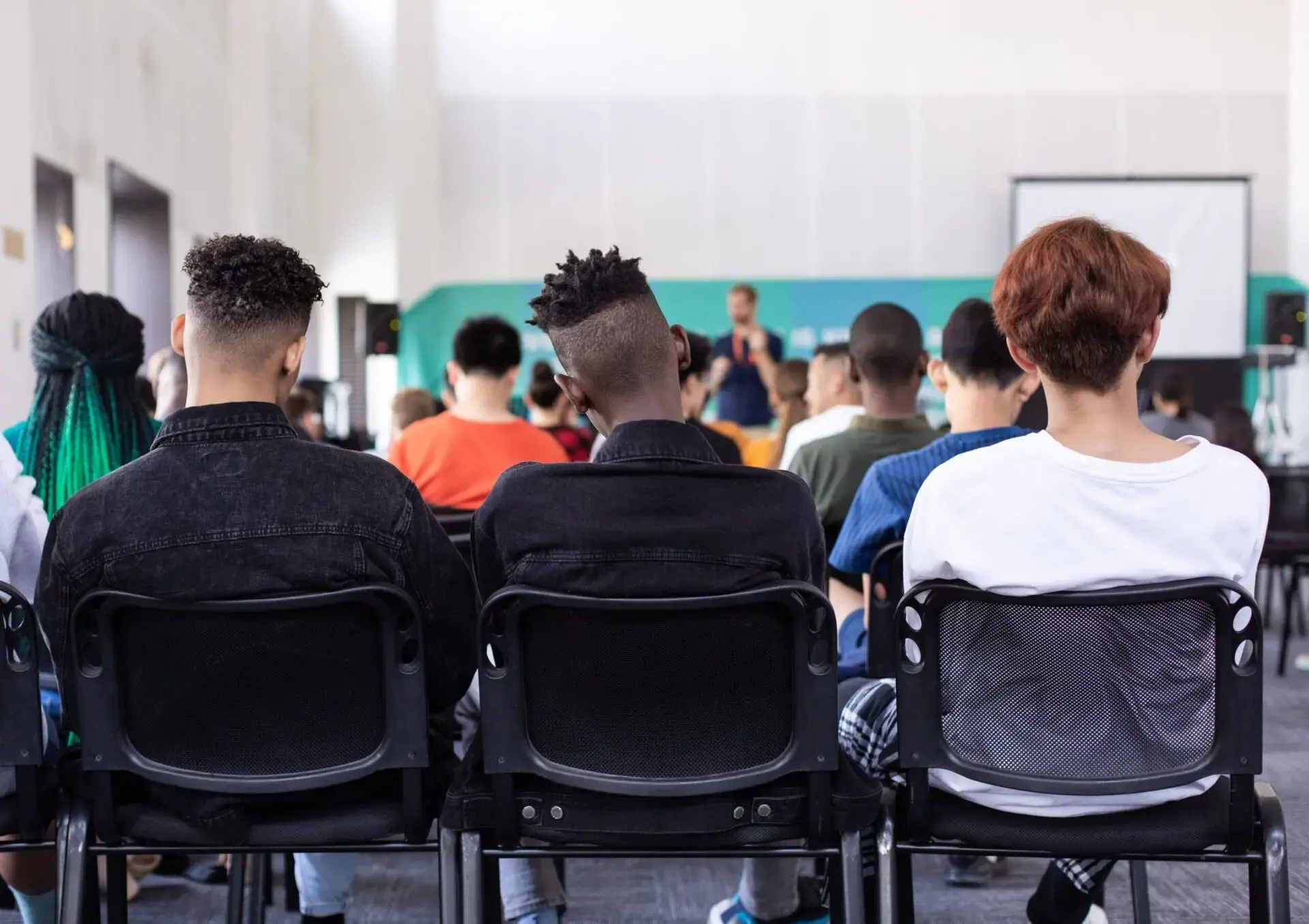A group of people sit in rows of chairs, facing forward and listening to a speaker at the front of a bright room with a projector screen. The focus is on the backs of three individuals in the foreground.