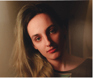 A person with long, light brown hair and fair skin looks at the camera with a neutral expression, softly lit against a blurred, warm-toned background.