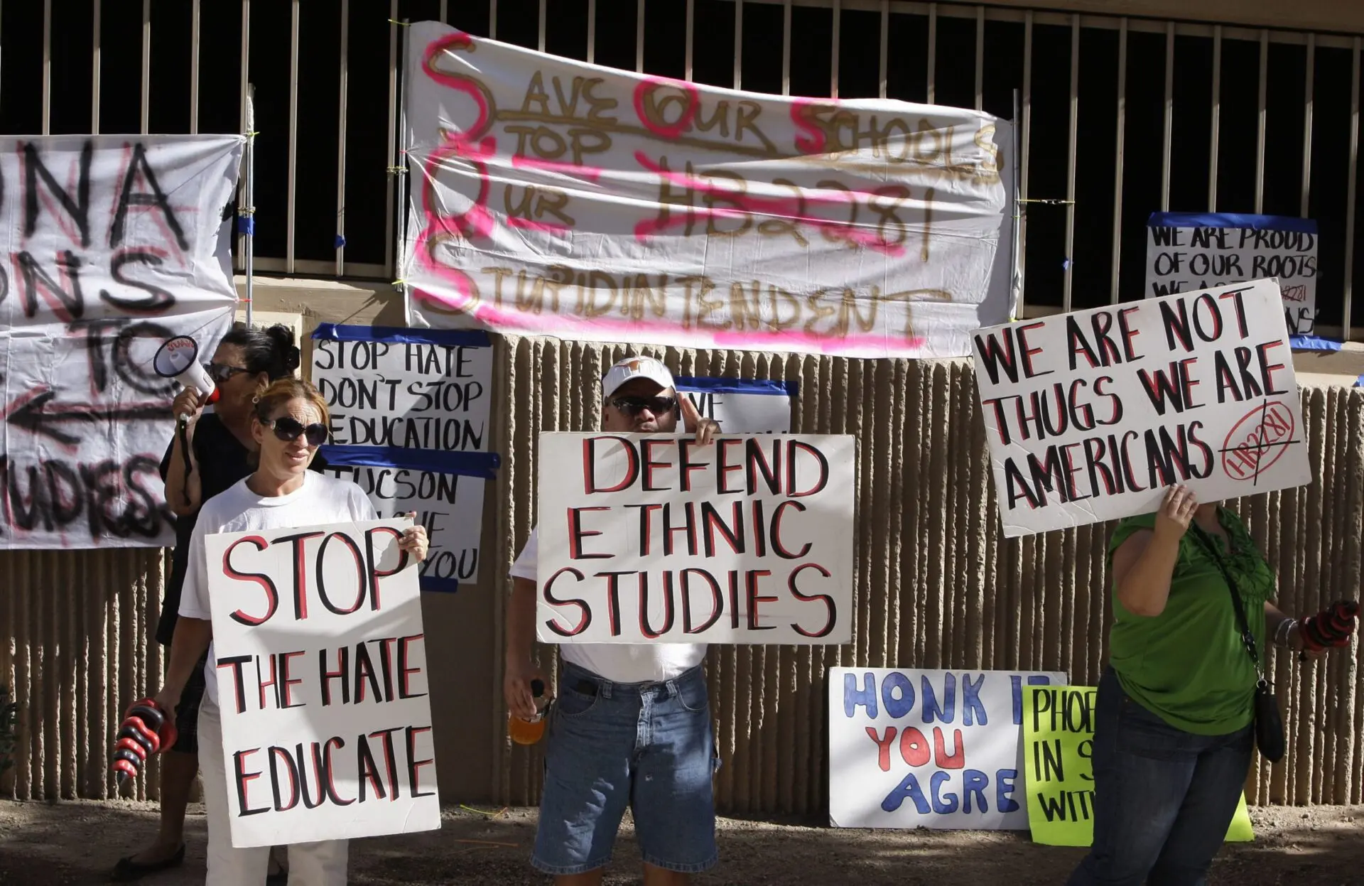 Three people hold protest signs supporting ethnic studies and education. Other signs and banners about ethnic studies, American identity, and opposing educational gag orders or bans are visible in the background, attached to a wall and railings.