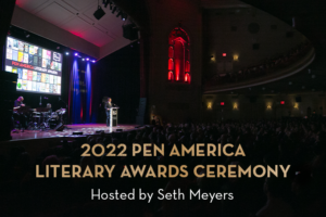 A person stands at a podium on stage during the 2022 PEN America Literary Awards Ceremony, hosted by Seth Meyers, with a large audience and colorful lights illuminating the venue.