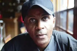 A man wearing a dark baseball cap and shirt sits indoors by a window, looking directly at the camera with a neutral expression.