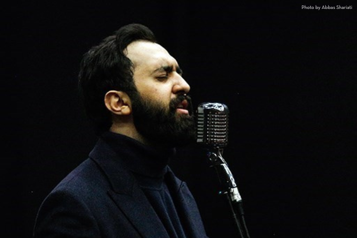 A man with a beard and dark hair sings passionately into a vintage microphone against a black background. He is wearing a dark turtleneck and blazer.
