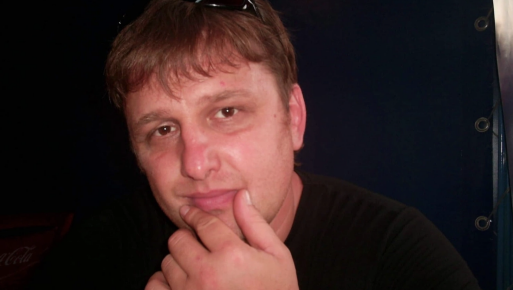 A man with light brown hair rests his chin on his hand and looks at the camera. He is wearing a black shirt and sunglasses on his head, with a dark background behind him.