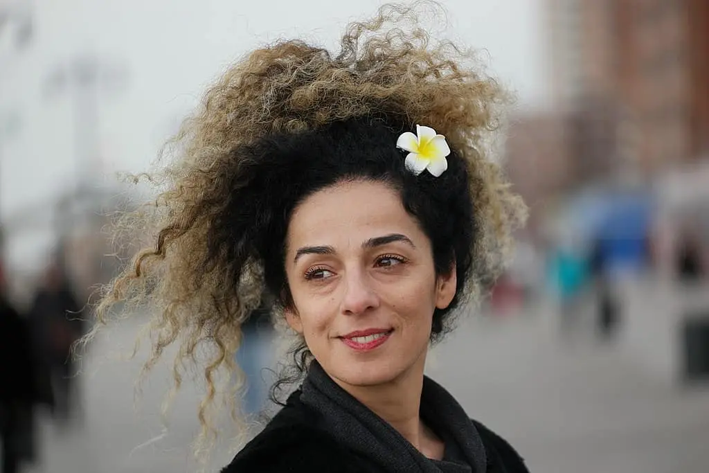 A woman with curly hair tied up, wearing a white and yellow flower in her hair, smiles slightly while standing outdoors. The background is blurred with people and buildings visible.