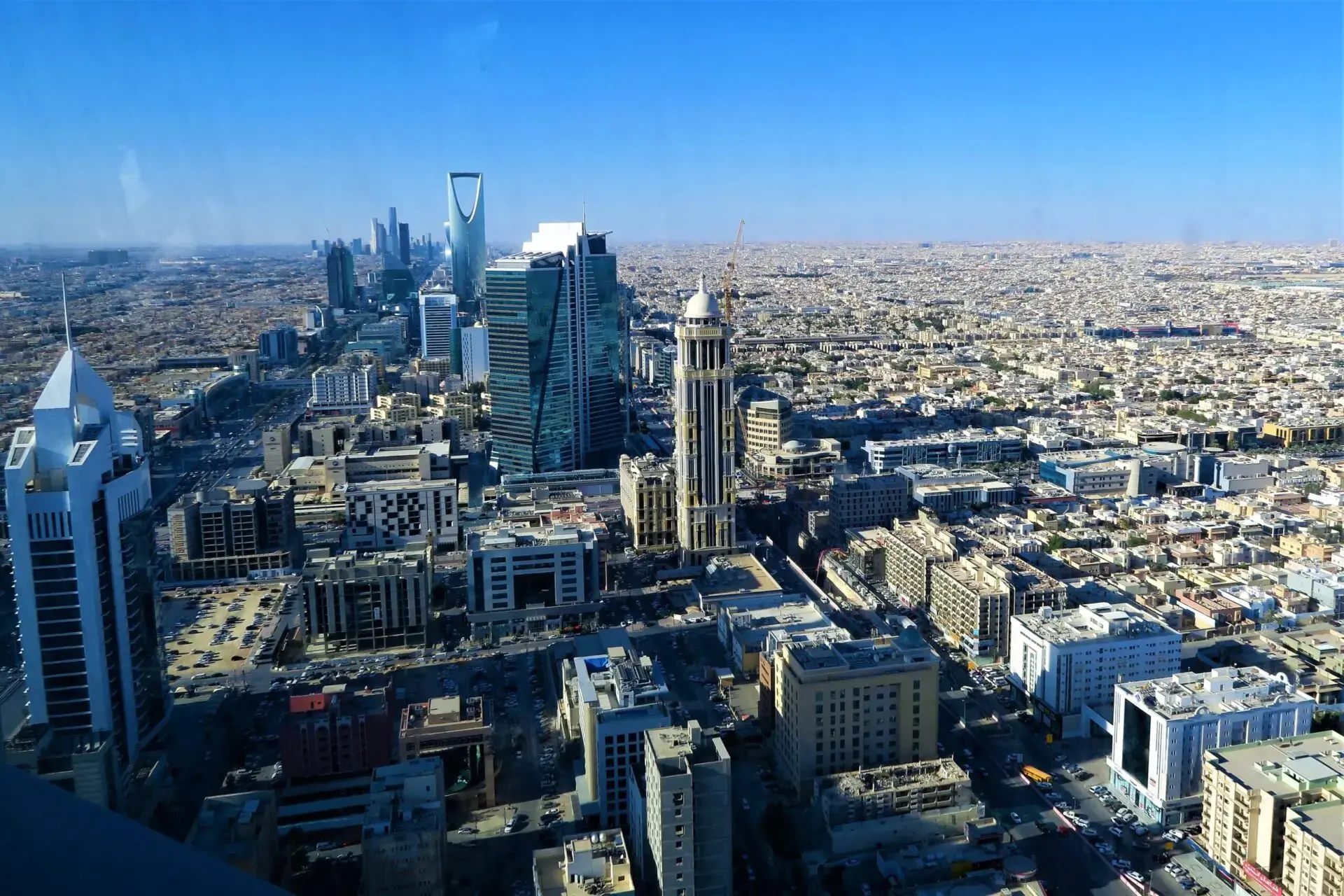 Aerial view of Riyadh, Saudi Arabia, featuring modern skyscrapers, including the iconic Kingdom Centre, surrounded by dense urban buildings under a clear blue sky.