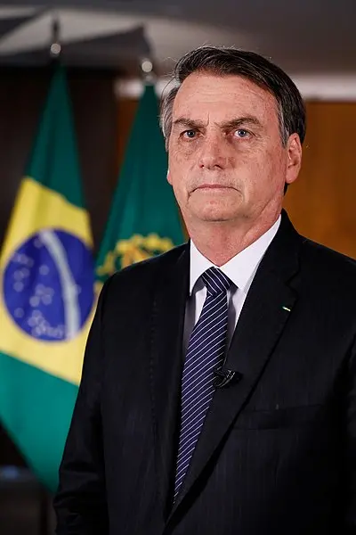 A man in a black suit and striped tie stands in front of two Brazilian flags, looking directly at the camera with a neutral expression.