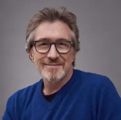 A middle-aged man with gray hair, a short beard, and glasses, wearing a blue sweater and smiling in front of a plain gray background.