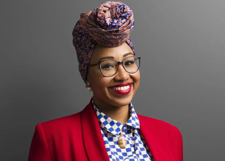 A smiling woman wearing glasses, a colorful patterned headwrap, red blazer, and geometric-print shirt stands against a plain gray background.