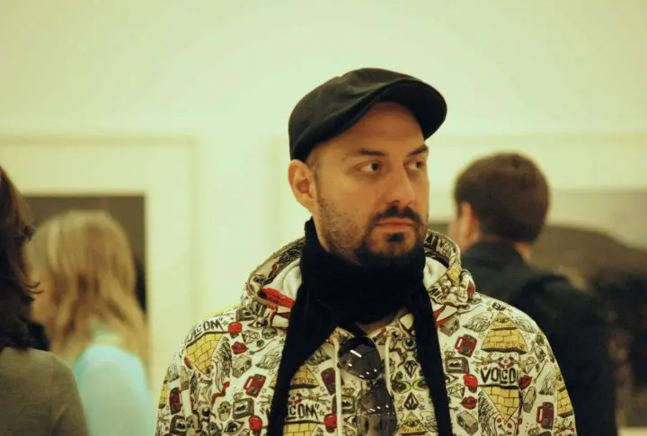 A bearded man wearing a patterned hoodie, black scarf, and black cap stands indoors with a neutral expression. Sunglasses hang from his hoodie. Blurred people and artwork are visible in the background.