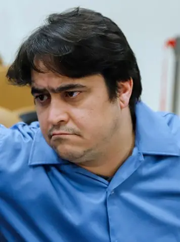 A man with dark hair and a serious expression wears a blue collared shirt, looking slightly downward and to the side. The background is out of focus.