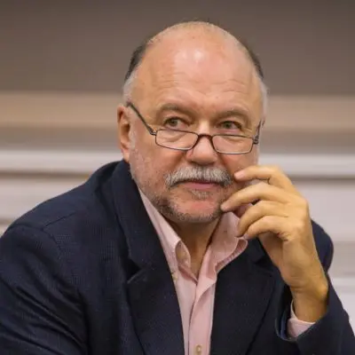 An older man with glasses, a mustache, and a beard is wearing a navy blazer and a light pink shirt. He is sitting with his hand touching his face, looking thoughtful.