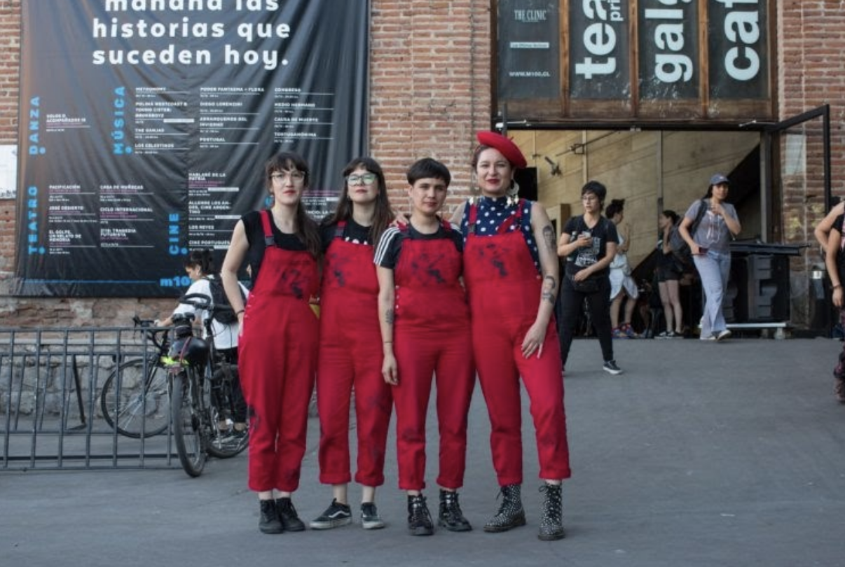 Four people wearing matching red overalls and boots stand together outdoors, posing for a photo. They are in front of a brick building with large posters and other people in the background.