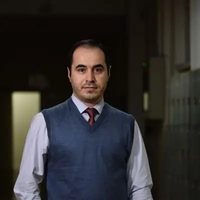 A man with short dark hair, wearing a white shirt, a red tie, and a blue sweater vest, stands indoors in a dimly lit hallway, facing the camera with a neutral expression.