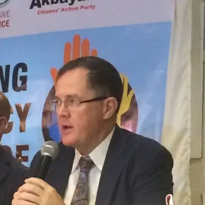 A man in a suit and tie speaks into a microphone at an event, with a blue and orange banner and logos visible in the background.