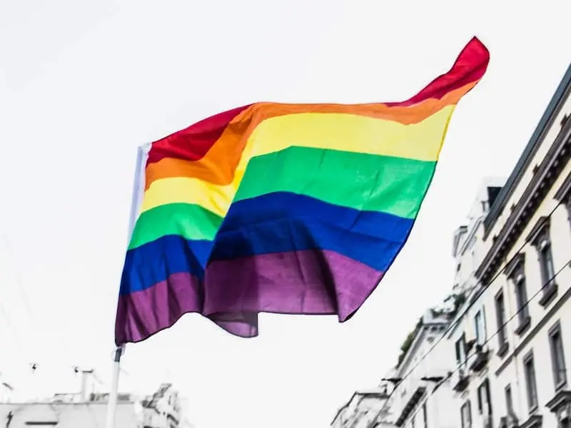 A rainbow pride flag waves in the air against a bright sky, with the tops of buildings visible on either side.