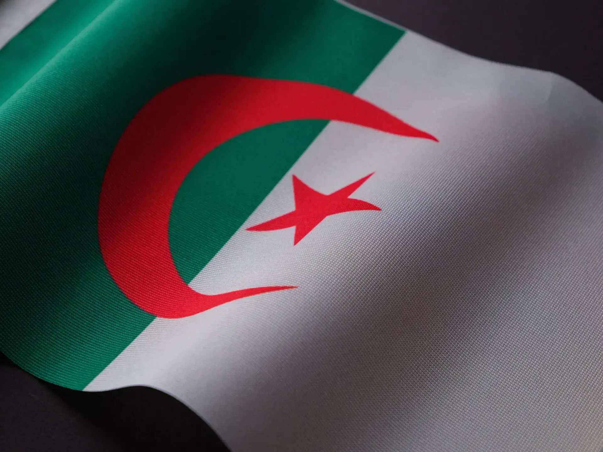 A close-up of the Algerian flag, featuring a green and white vertical split with a red crescent and star in the center. The fabric texture and folds are visible.