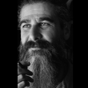 Black and white photo of a bearded person with long hair, holding something near their mouth, looking slightly upward, and smiling softly. The lighting highlights their face, creating a contemplative mood.