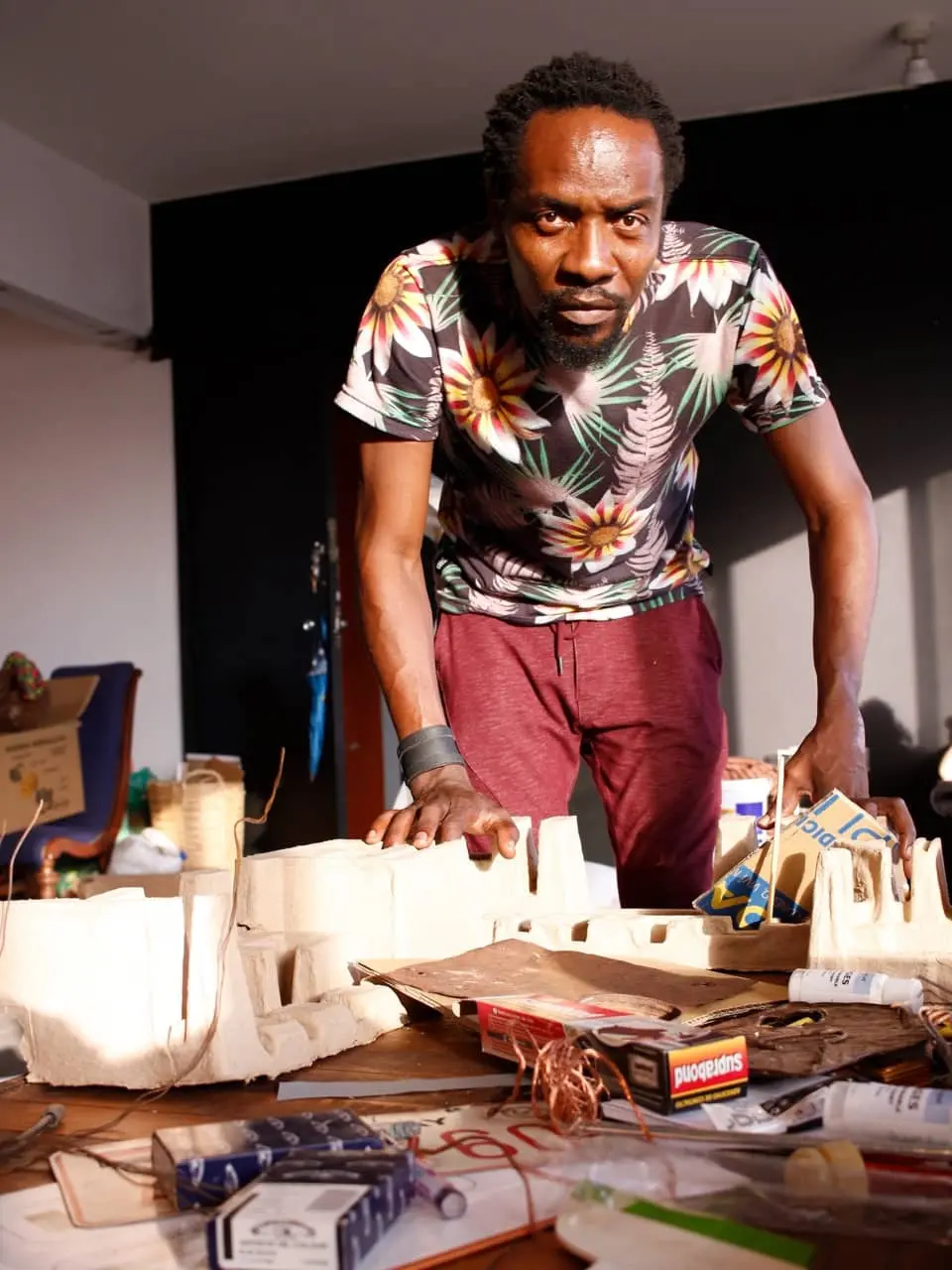 A man in a floral shirt and maroon pants leans over a table covered with art supplies and cardboard pieces, looking directly at the camera in a well-lit room.