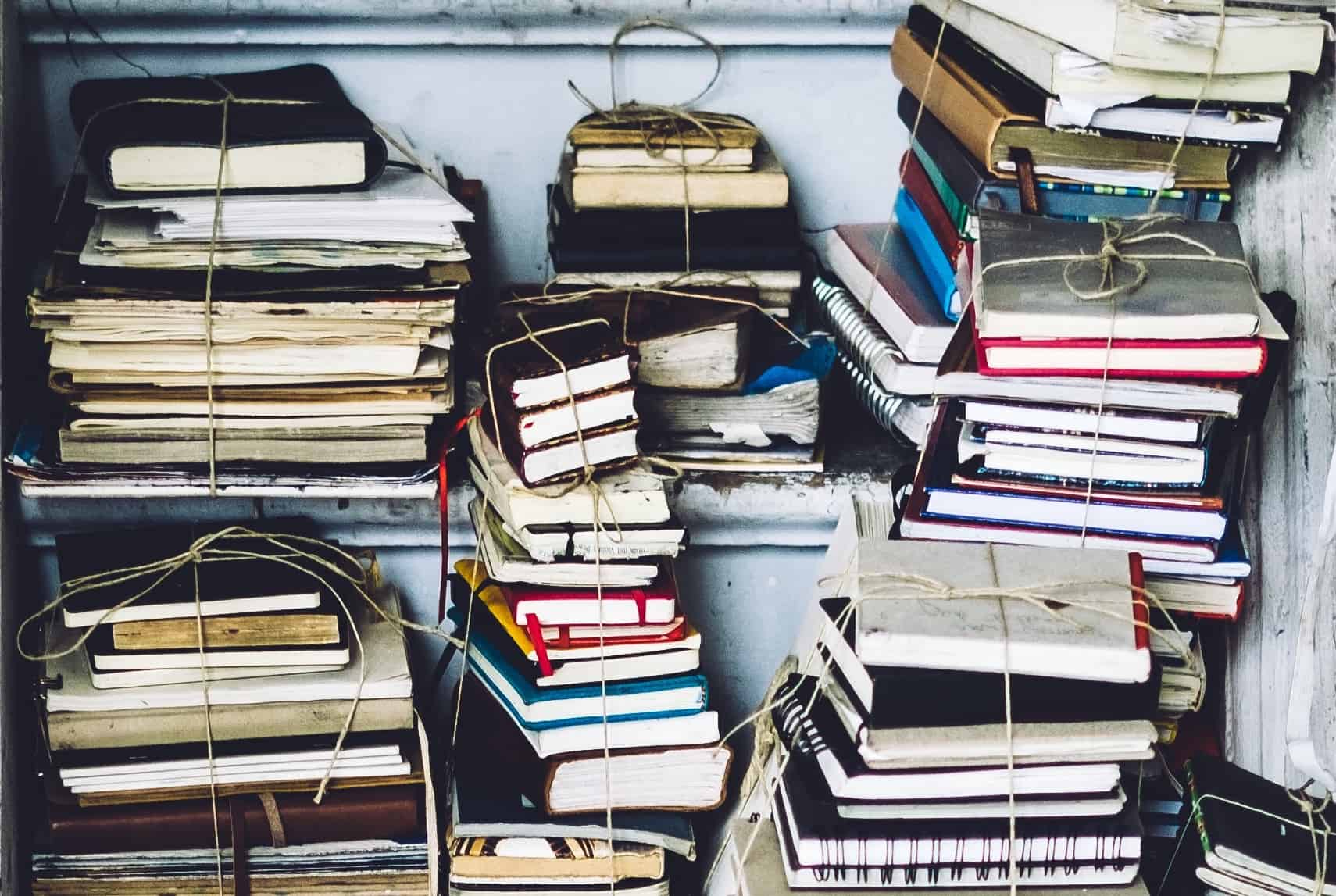 Stacks of assorted old books and notebooks tied with string are piled on shelves, creating a cluttered and vintage appearance. The books vary in size, color, and condition.