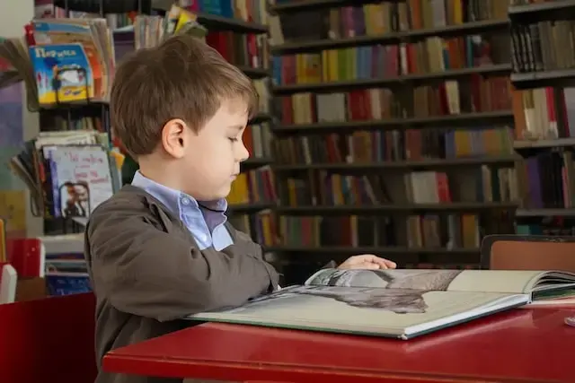 A young boy sits at a red table in a library, looking at a large open book. Shelves filled with colorful books are visible in the background.