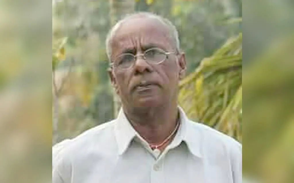 An older man with short gray hair and glasses is wearing a light-colored button-up shirt, standing outdoors with greenery and tree branches in the background.