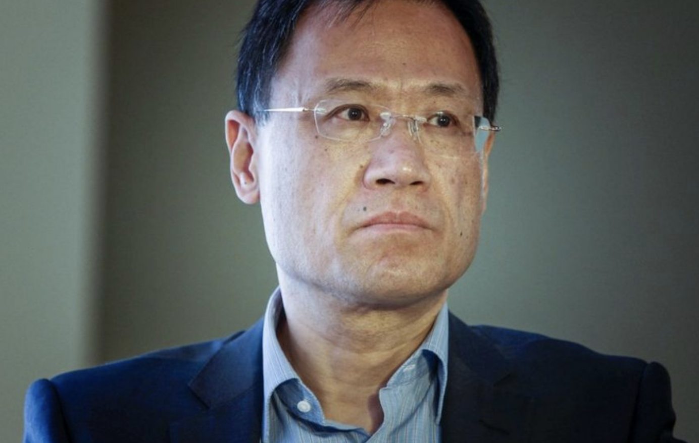 A man wearing glasses and a blue suit jacket over a light blue shirt looks serious while facing slightly to the left, with a neutral background behind him.