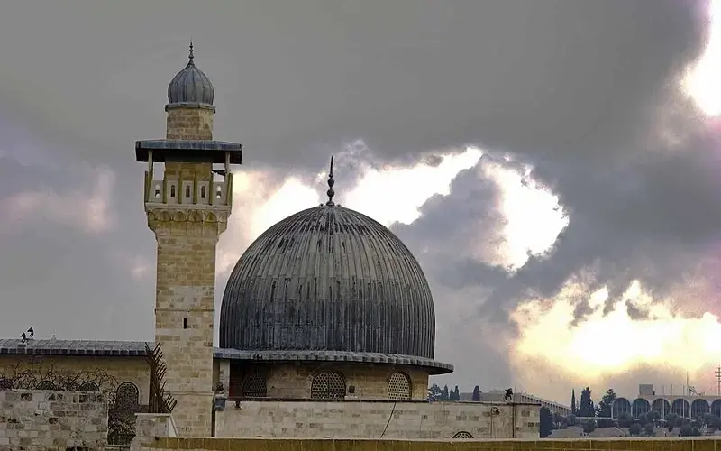A mosque with a large grey dome and a single minaret stands against a dramatic cloudy sky at sunset, with sunlight breaking through the clouds.