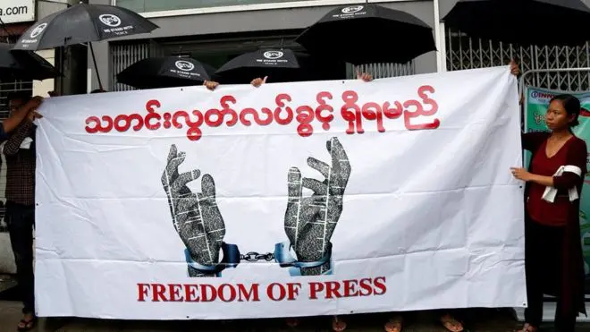 People hold a banner with handcuffed hands breaking free, text in Burmese, and FREEDOM OF PRESS in English, during a protest. Some protesters hold black umbrellas with printed white logos.