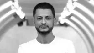 A man with short dark hair and a trimmed beard, wearing a plain white shirt, stands in front of a blurred, brightly lit tunnel-like background.