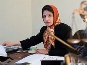 A woman wearing a patterned headscarf and black jacket sits at a desk with papers, a teacup, and office items, looking toward the camera in an indoor office setting.