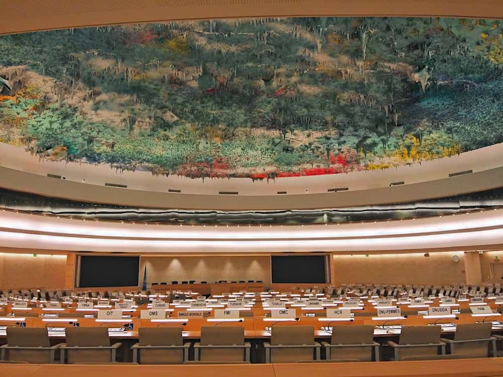 A large, modern conference room with rows of desks and chairs, each desk labeled with country names, and a vibrant, colorful abstract ceiling mural overhead. The room is empty and well-lit.