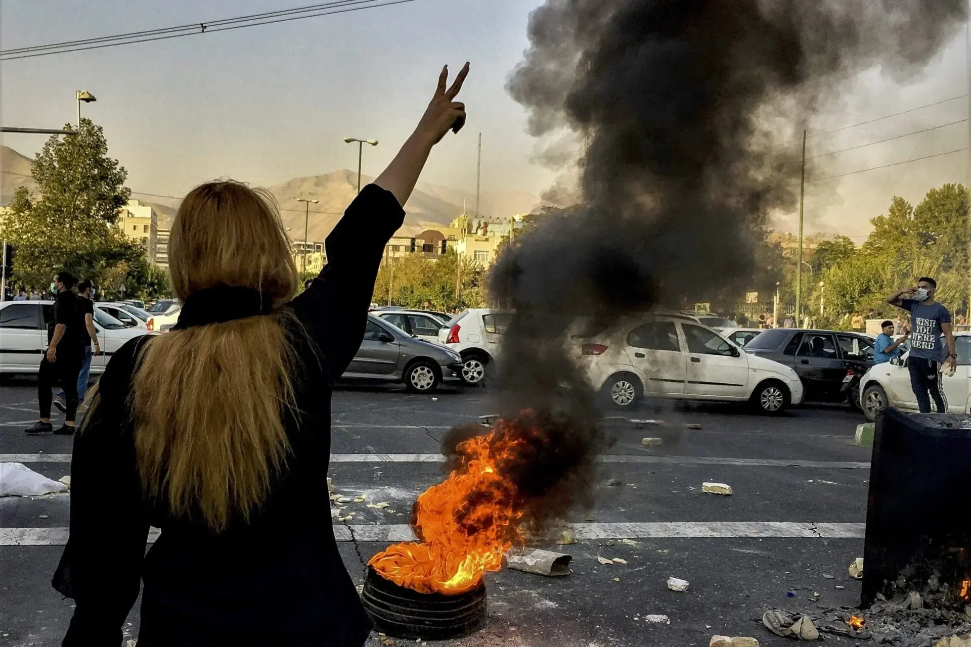 A person with long blonde hair raises a peace sign near a burning tire, with thick black smoke rising. Cars and people are in the background on a city street during daylight.