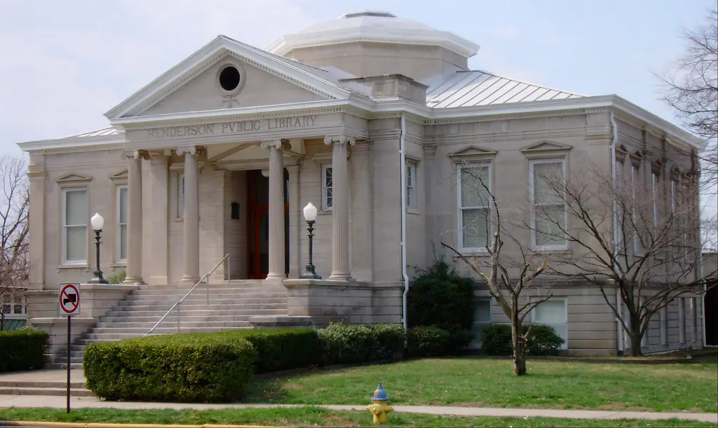 A historic stone building with columns and a pediment, labeled “Henderson Public Library,” stands amidst bushes and trees, with steps leading to the entrance and a fire hydrant in front.