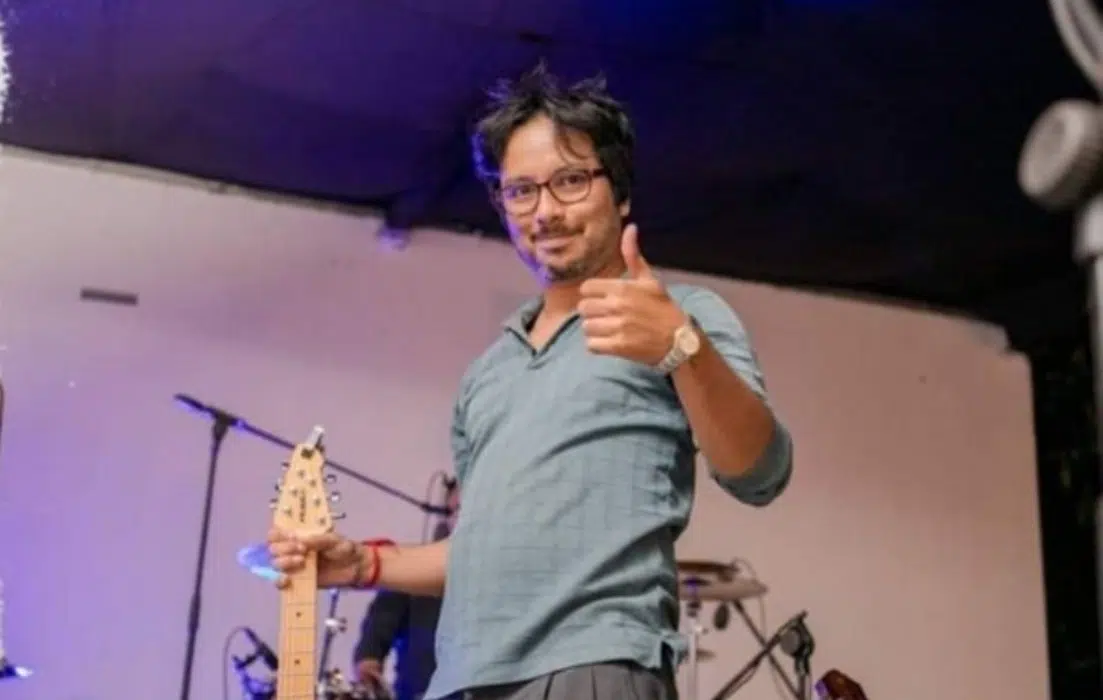 A man wearing glasses and a blue shirt stands on a stage with a guitar in one hand and gives a thumbs up with the other. Musical instruments and equipment are visible in the background.