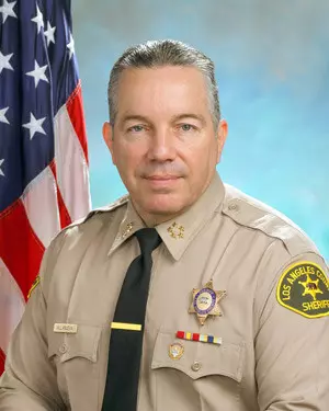 A man in a tan Los Angeles County Sheriffs uniform poses for an official portrait, sitting in front of a U.S. flag with a blue background. He has short gray hair and wears a black tie and several badges.