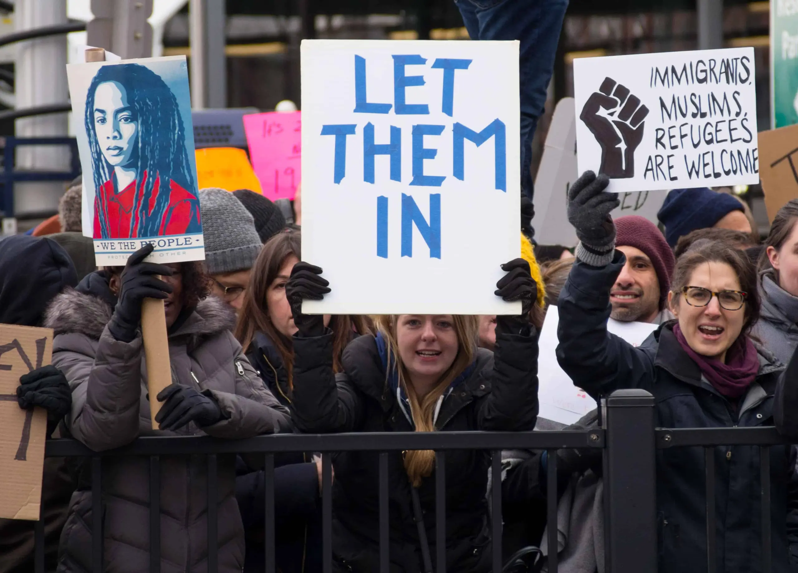 A diverse group of protesters hold signs, including LET THEM IN, IMMIGRANTS MUSLIMS REFUGEES ARE WELCOME with a raised fist, and an illustrated portrait sign, during a public demonstration.