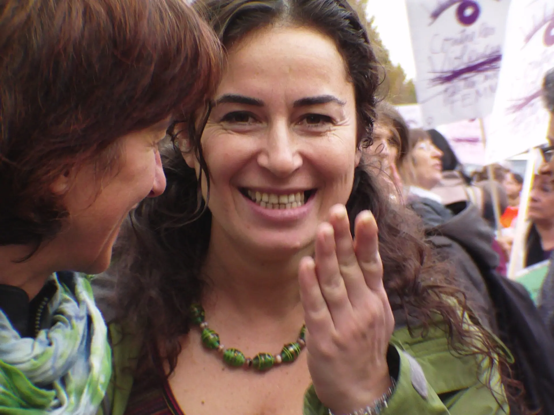 A smiling woman with long curly hair and a green necklace holds her hand near her face while talking to another woman in a crowd at an outdoor event or protest. Signs can be seen in the background.