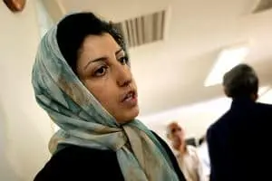 A woman wearing a light-colored headscarf stands indoors, looking to the side. The background is softly lit with other people visible but out of focus.