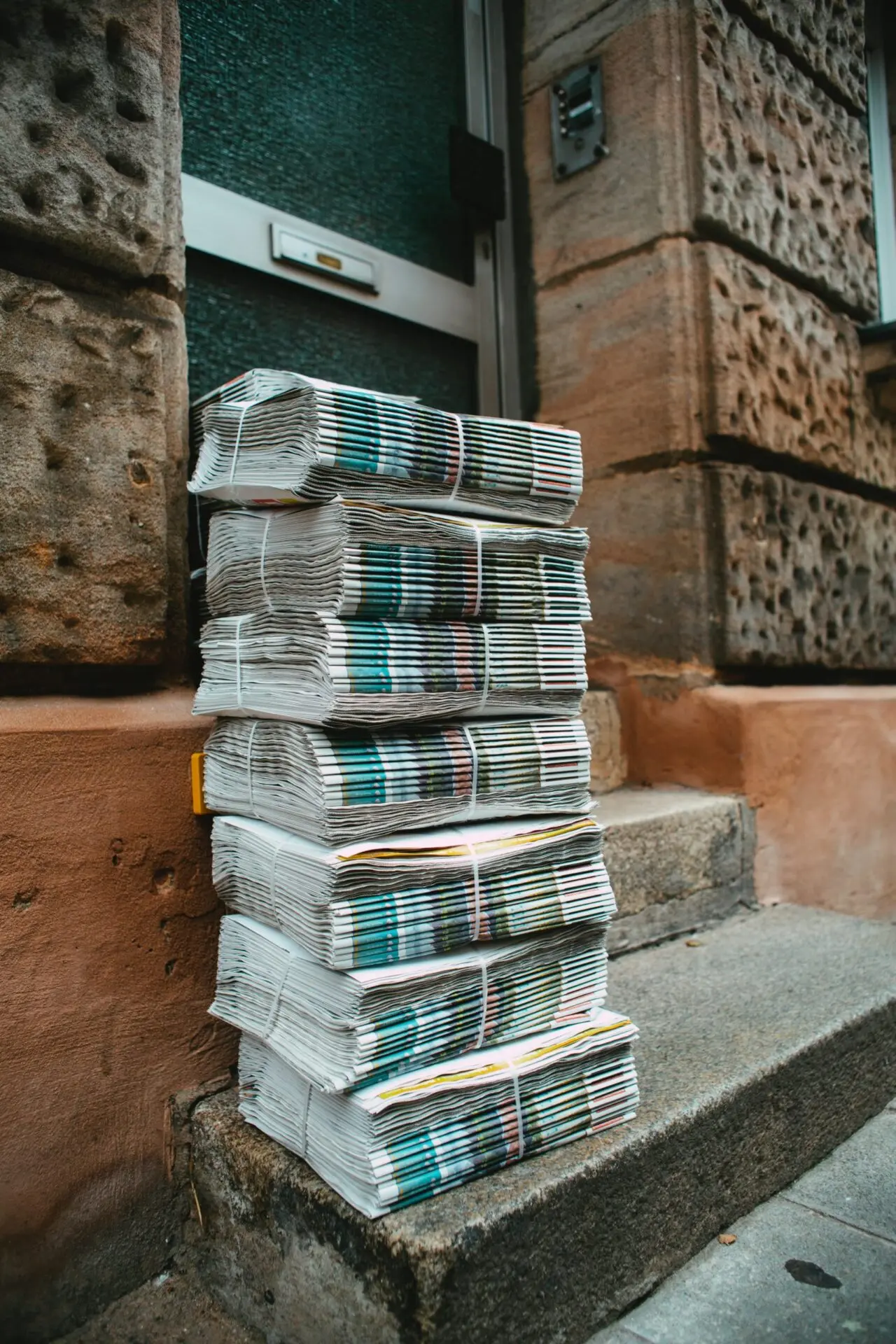 A stack of bundled newspapers sits on a stone doorstep outside a building with textured, brown brick walls.