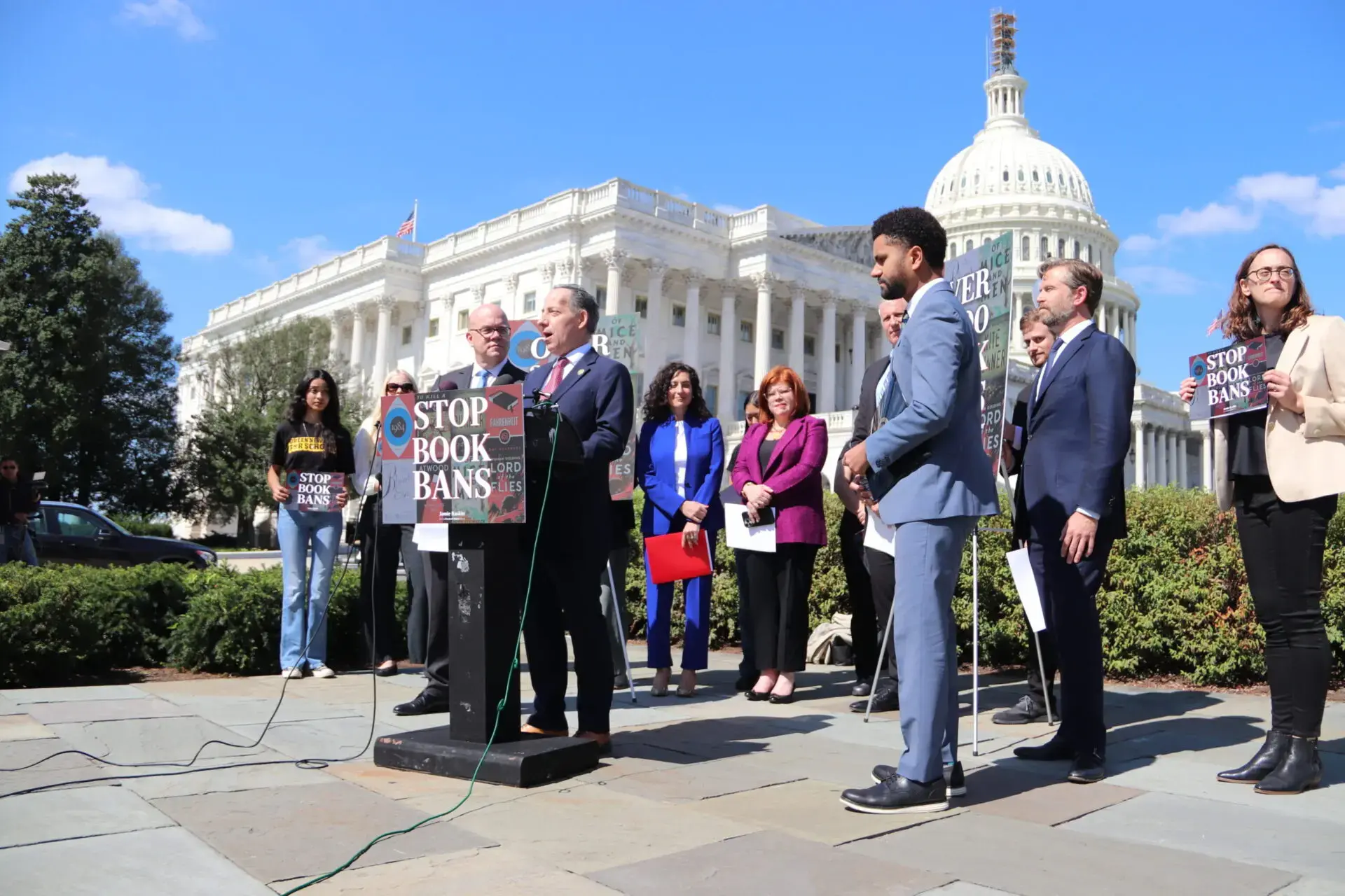 A group of people stand outside the U.S. Capitol holding a press conference. A speaker at a podium displays a sign reading STOP BOOK BANS, while others listen or hold signs in the background.