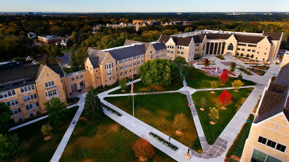 Aerial view of a university campus with stone buildings, green lawns, pathways, flagpole, and colorful trees, set against a backdrop of trees and distant cityscape.