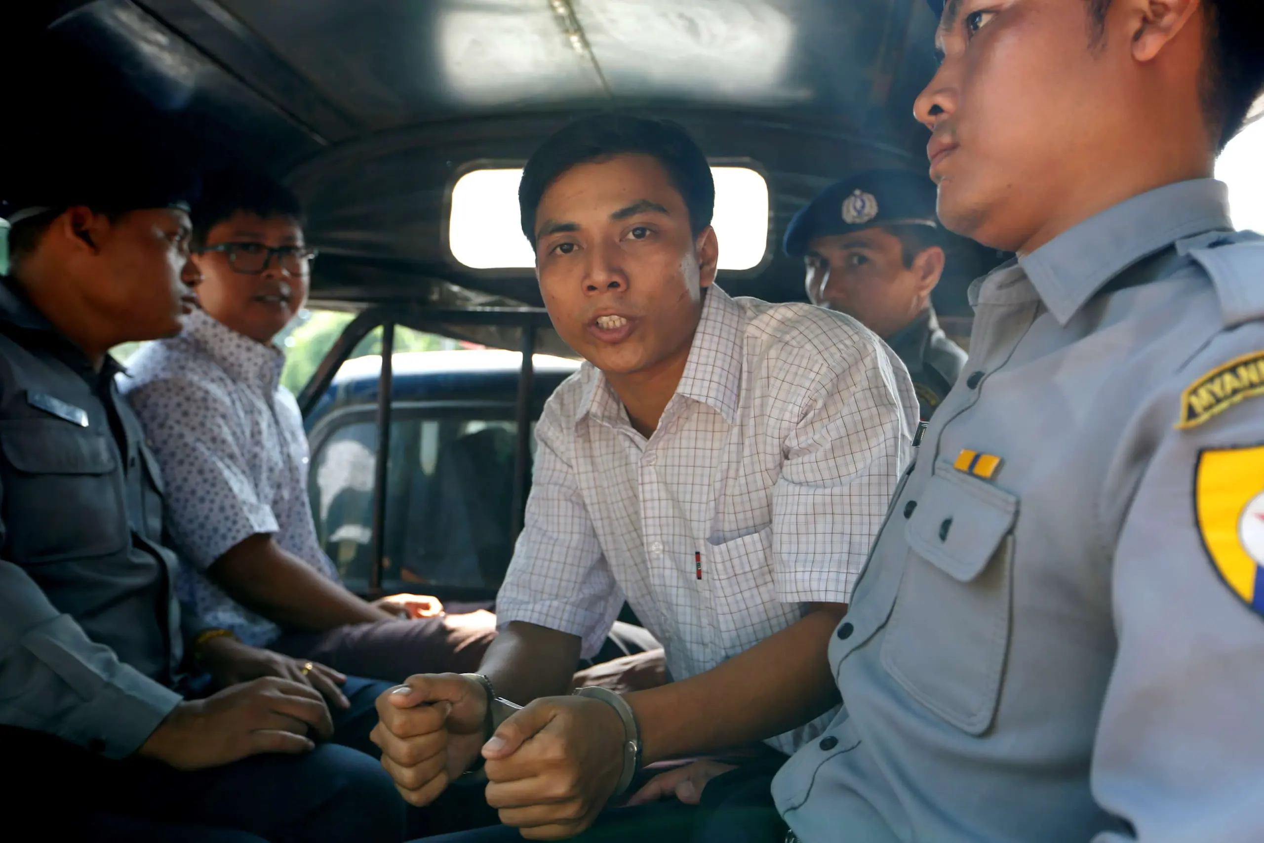 A man in handcuffs, possibly one of the Reuters journalists, sits between police officers and another man in the back of a vehicle, looking directly at the camera with a serious expression. The officers wear uniforms and caps.