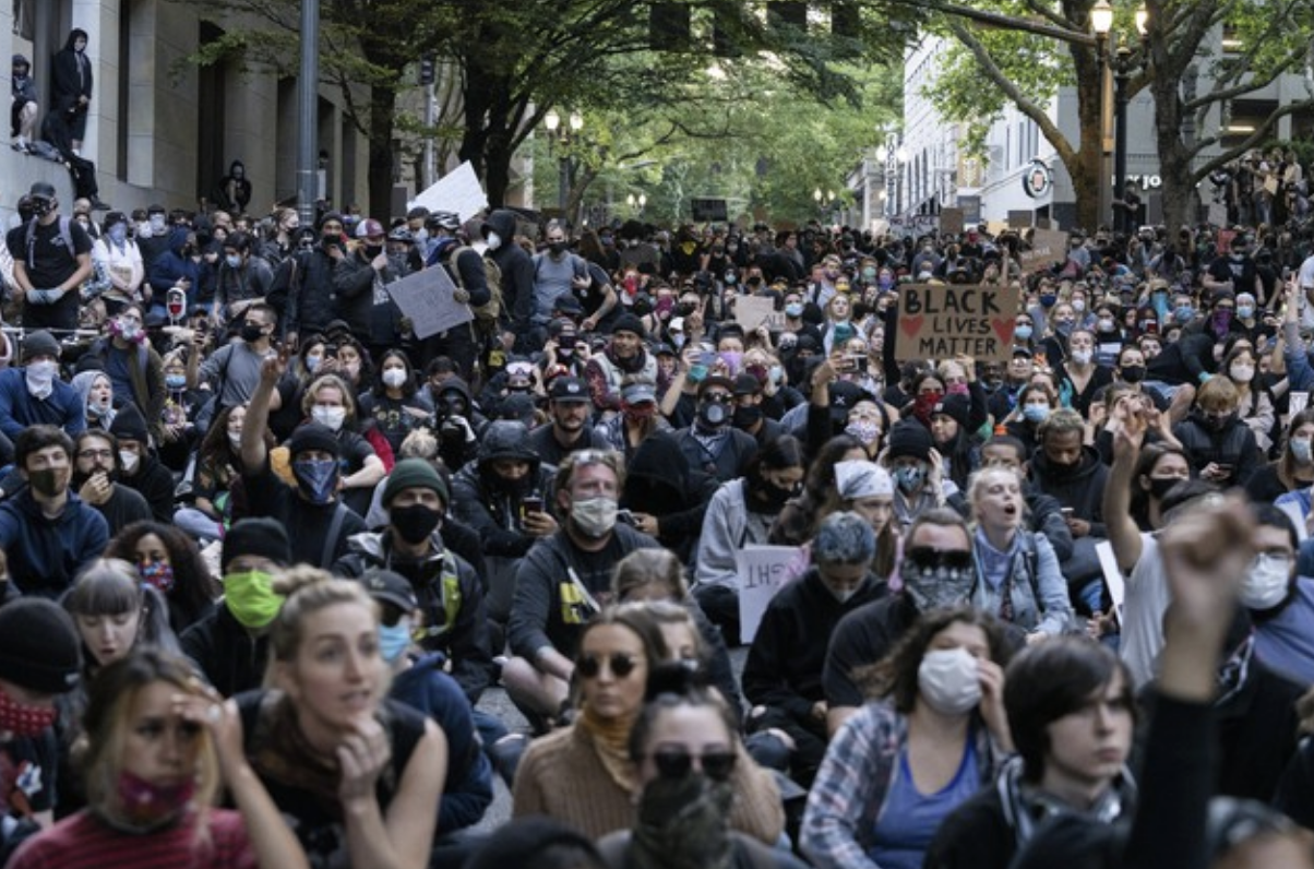 A large crowd of people, many wearing masks, sit closely together outdoors holding protest signs, including one that reads Black Lives Matter, surrounded by trees and city buildings.