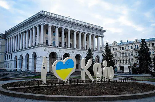 A large sign reading I ♥ Kyiv with a heart in the colors of the Ukrainian flag stands in front of a grand, white neoclassical building with columns in Kyiv, Ukraine, under a cloudy sky.