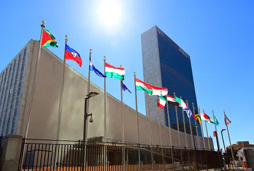 Flags of various countries fly outside the United Nations headquarters in New York City on a sunny day, with the UN building and a clear blue sky in the background.